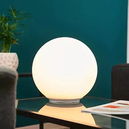 Darc — Lampe sphérique blanche posée sur une table transparente, éclairant faiblement une pièce aux murs teintés de bleu-vert, avec une chaise grise e