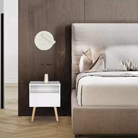 Darc — Une lampe murale circulaire blanche fixée sur un mur en bois sombre, éclairant une petite table de chevet blanche à tiroirs avec pieds en bois