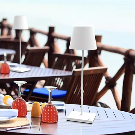 Darc — Une table en bois sur une terrasse avec deux lampes blanches à abat-jour, des verres à vin rouges posés sur des supports, des coussins colorés