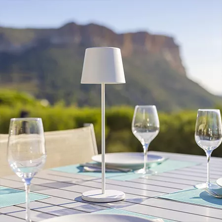 Darc — Une table extérieure en carrelage bleu clair avec une lampe à lumière douce blanche au centre, entourée de deux verres à vin vides, le tout fac
