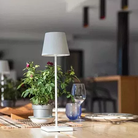 Darc — Une lampe design blanche en forme de cone posée sur une table en bois clair, entourée d'une plante verte en pot et d'un verre à vin bleu transp
