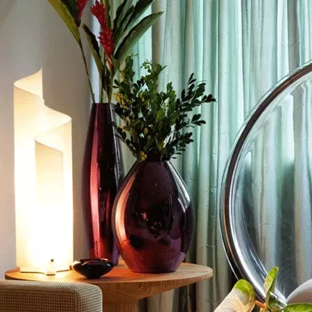 Darc — Une table basse en bois clair sur laquelle reposent deux vases en verre teinté, l’un rouge et l’autre violet foncé, remplis de feuillages verts