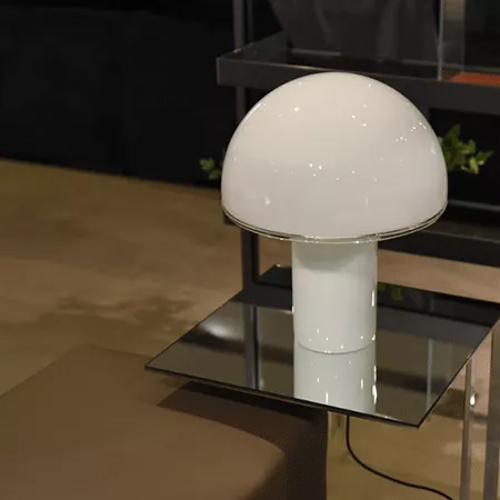 Darc — Lampe design en forme de champignon blanc posée sur une table basse noire et transparente, avec une lumière douce émise par son sommet arrondi.