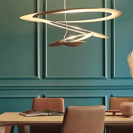 Darc — Une suspension lumineuse dorée en forme d’anneaux suspendus au plafond, éclairant une table basse avec deux fauteuils en cuir brun et un livre