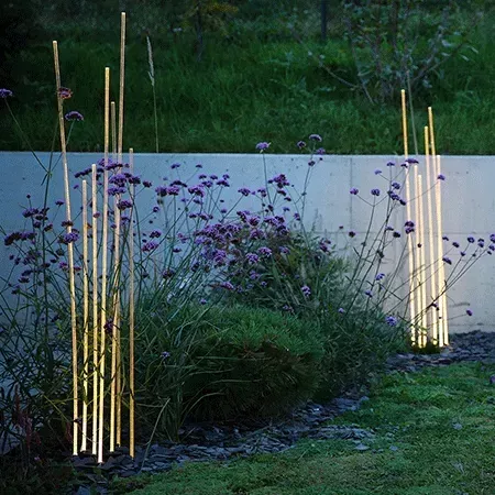 Darc — Quatre luminaires fins et élancés en métal doré, plantés verticalement dans un massif de fleurs violettes et de végétation verte, éclairés en s