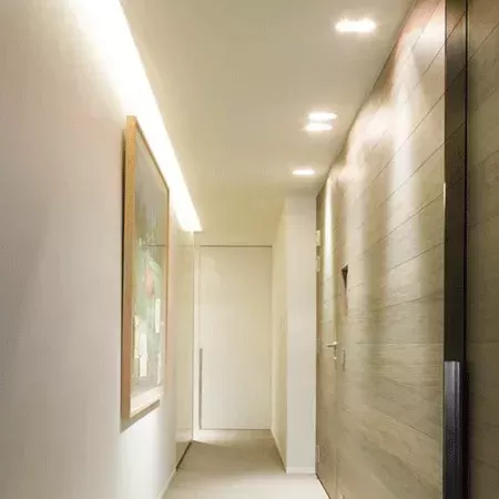 Darc — Un couloir étroit menant à une salle de bain moderne, avec des murs blancs, un miroir encadré de bois clair à gauche, un éclairage indirect au