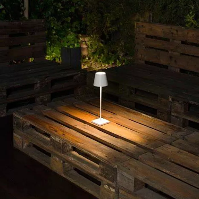 Darc — Une lampe blanche moderne posée sur une palette en bois, éclairant légèrement une zone extérieure avec une ambiance chaleureuse et cosy