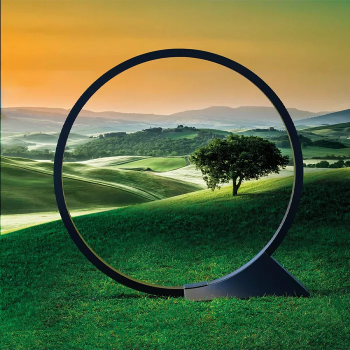 Darc — Un cercle noir métallique posé sur une prairie verte encadre une vue panoramique de collines verdoyantes, d’un arbre isolé et d’un coucher de s