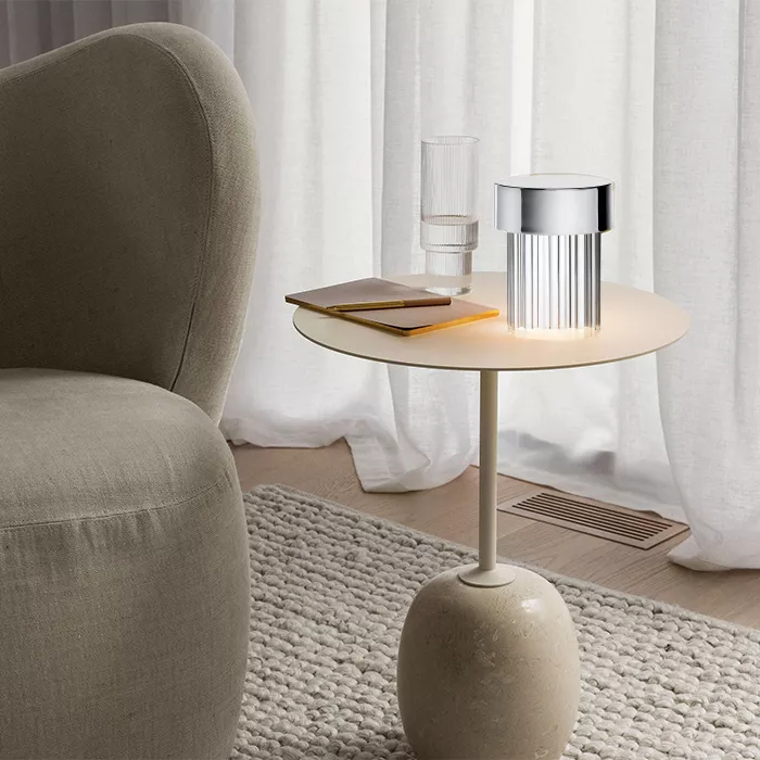 Darc — Une table basse ronde en bois clair posée sur un pied en pierre beige, éclairée par une lampe design métallique à lumière douce. À côté, un ver