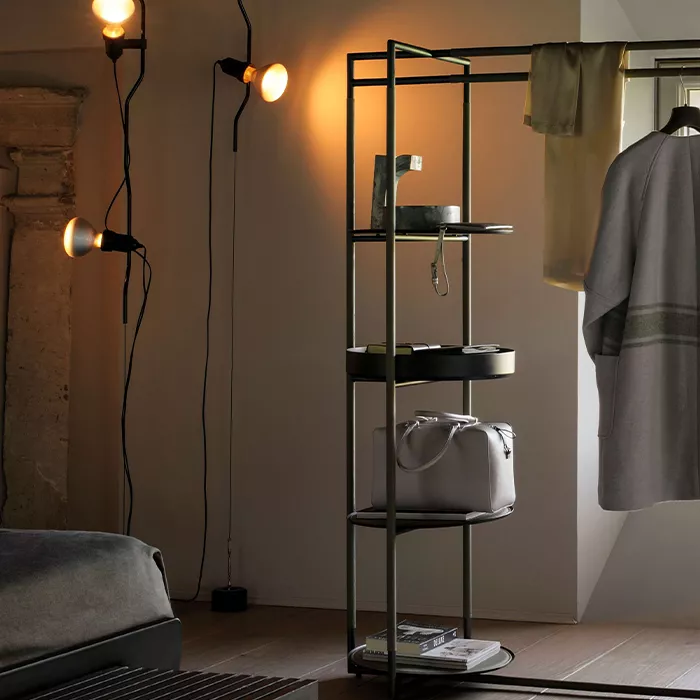 Darc — Une armoire métallique noire à plusieurs étagères, éclairée par deux appliques murales, avec un sac posé sur une étagère et des vêtements suspe
