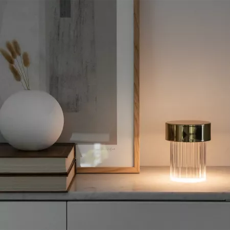 Darc — Une lampe design transparente avec un pied métallique doré posée sur un meuble blanc, éclairant légèrement une surface en bois clair. À côté, u