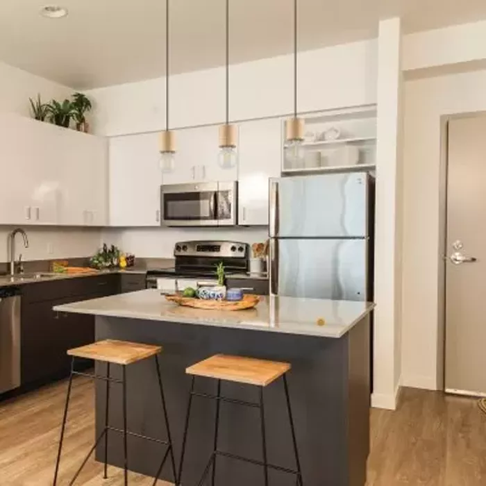 Darc — Une cuisine lumineuse équipée d’un îlot central en plan de travail blanc, entouré de deux tabourets en bois clair sur pieds métalliques noirs.