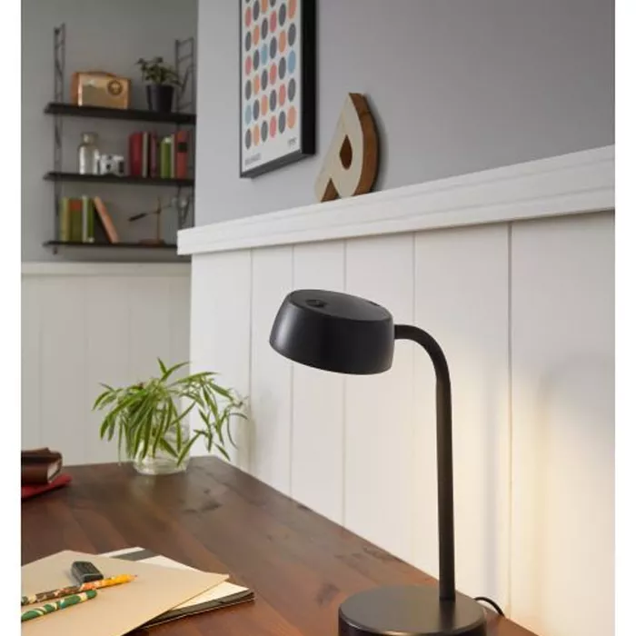 Darc — Une lampe de bureau noire et design, positionnée sur un bureau en bois avec une lumière douce dirigée vers l'avant, entourée d'une plante verte