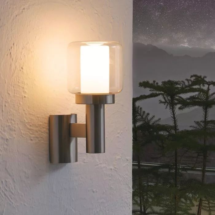 Darc — Une applique murale moderne en métal fixée sur un mur clair, émettant une lumière douce à travers un abat-jour en verre transparent. À côté, un