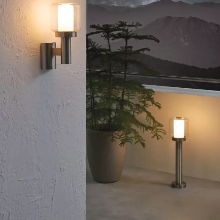 Darc — Deux luminaires extérieurs modernes éclairés : un applique mural fixé sur un mur blanc texturé et un lampadaire sol en métal avec abat-jour, en