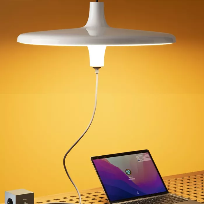 Darc — Une lampe suspendue blanche et design, fixée au plafond par un câble flexible, éclaire un bureau en bois clair. À côté, un ordinateur portable