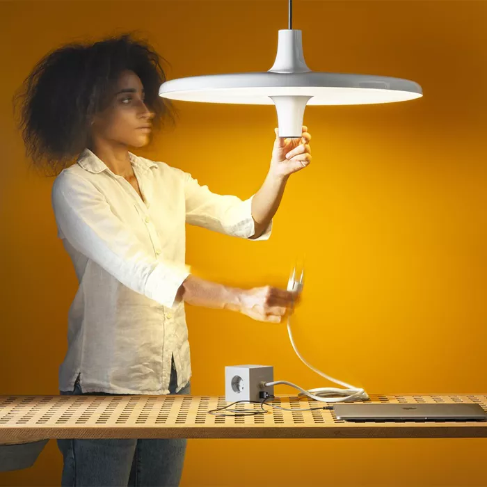 Darc — Une personne installe un luminaire design blanc suspendu au plafond, tenant le support tout en ajustant l’ampoule dans une pièce aux murs orang