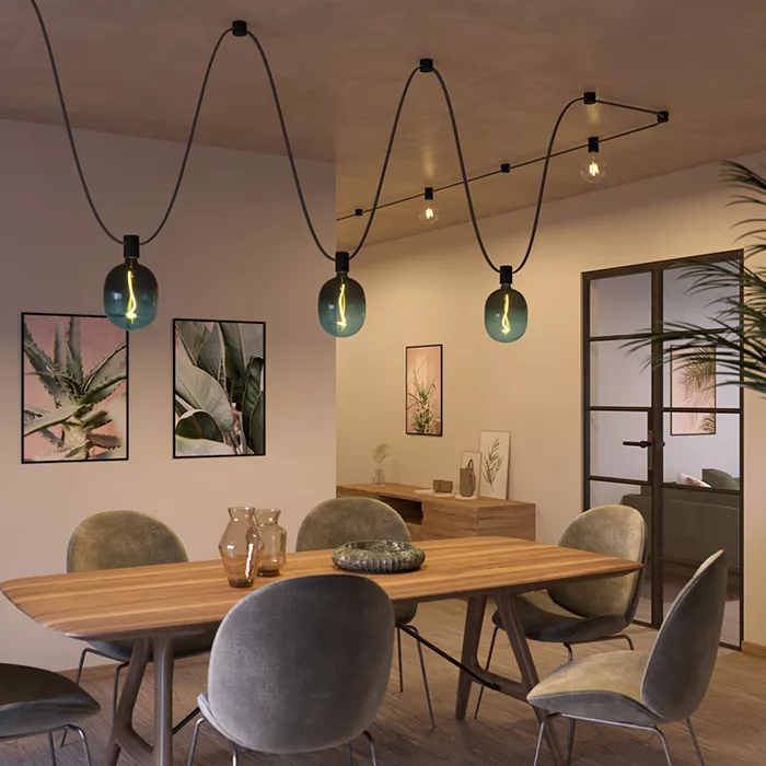 Darc — Une pièce lumineuse équipée d’une table en bois clair entourée de quatre chaises modernes. Trois lustres suspendus en forme de vagues, avec des