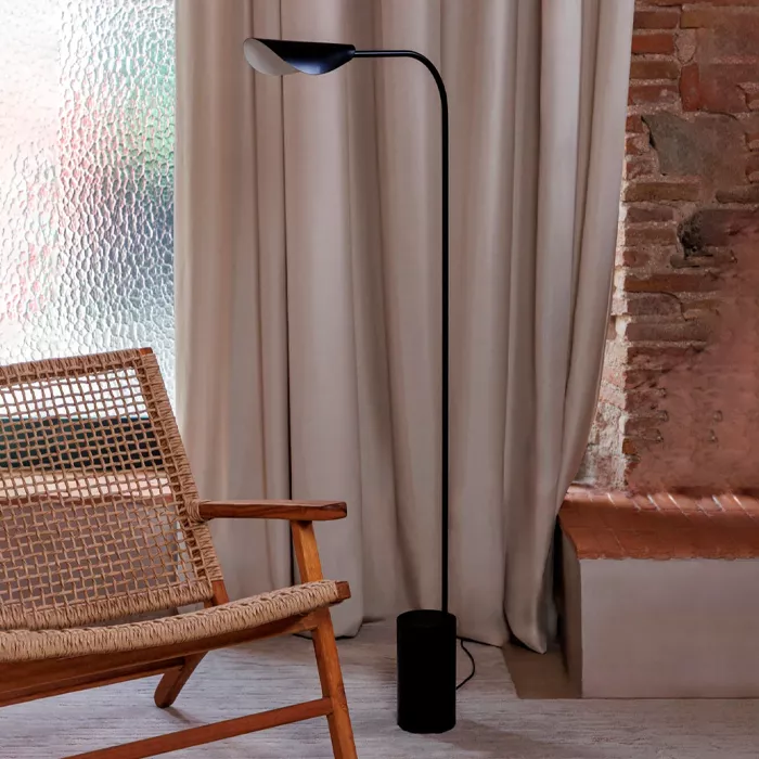 Darc — Un fauteuil en rotin naturel placé près d'une fenêtre voilée de rideaux beige clair, éclairé par une applique murale noire design en forme de c