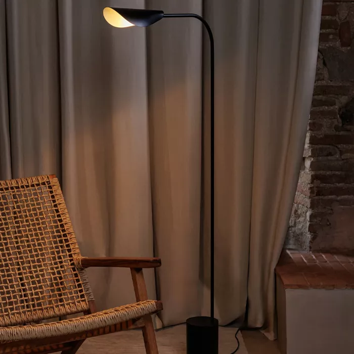 Darc — Une applique murale noire avec un abat-jour ovale éclairé est fixée près d’un rideau beige épais. À côté, un fauteuil en rotin avec une structu