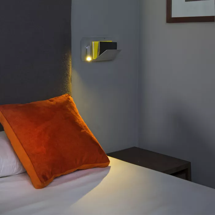 Darc — Une lampe murale design fixée près du lit, projetant une lumière douce et chaude sur un coussin orange et le drap blanc, dans une chambre aux m