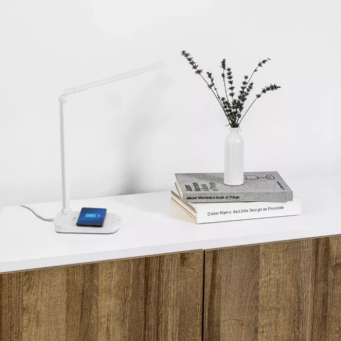 Darc — Un bureau épuré en bois clair avec une lampe LED flexible blanche allumée, un chargeur sans fil blanc et bleu, deux livres posés à plat dont un