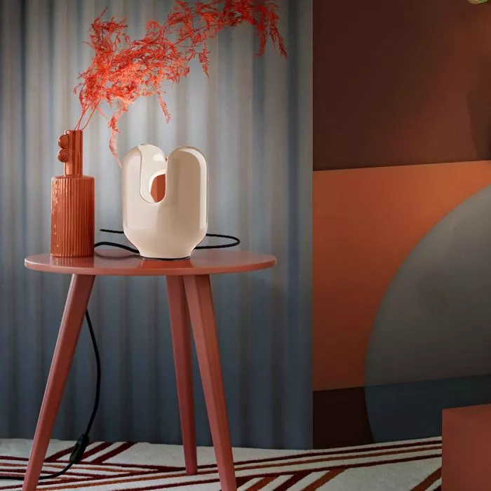 Darc — Une lampe de table design en forme de coquillage blanc posée sur un petit meuble rouge. À côté, un vase en terre cuite rouge contient une branc