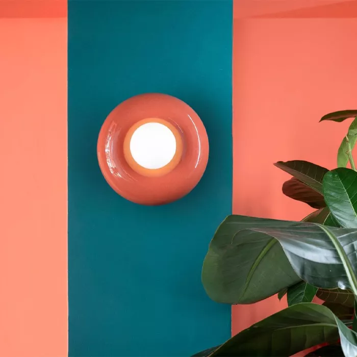 Darc — Un luminaire mural rond en céramique corail encastré dans un panneau bleu turquoise, éclairé par une ampoule douce, posé devant une plante vert