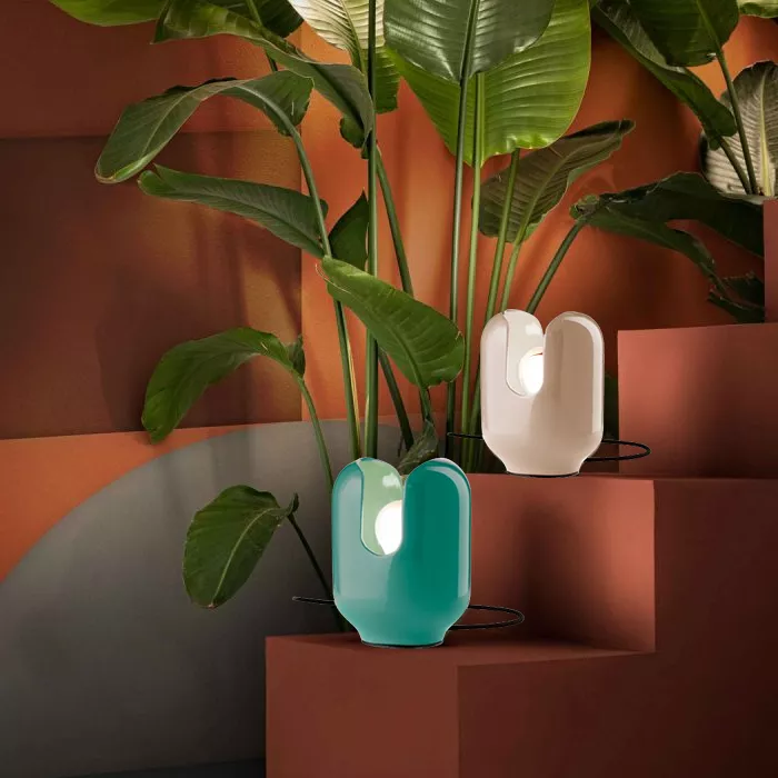 Darc — Deux luminaires en forme de vase, l’un turquoise et l’autre blanc, éclairés par une lumière douce, entourés de grandes feuilles vertes d’une pl