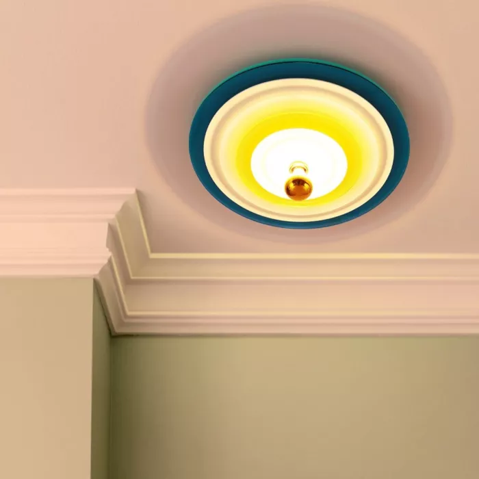 Darc — Une applique murale encastrée dans un plafond peint en rose pâle, entourée d’un cadre bleu clair, diffusant une lumière chaude et dorée.