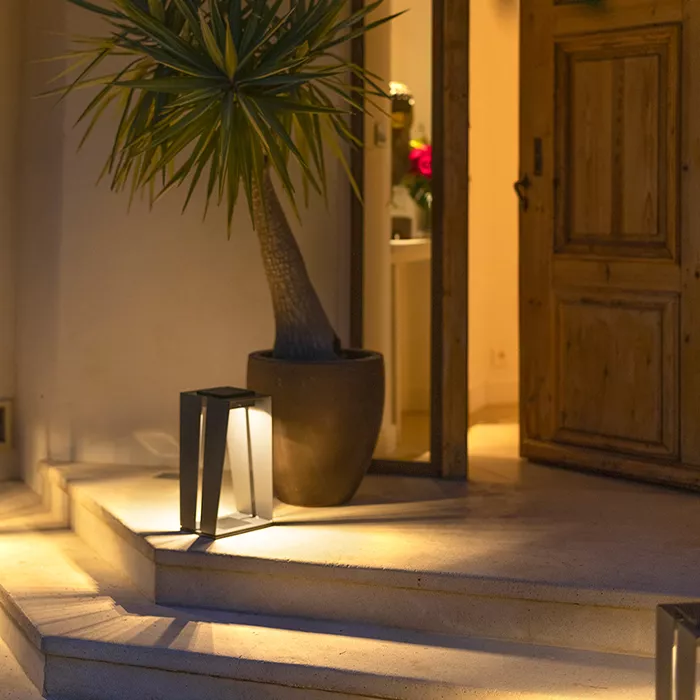 Darc — Un luminaire extérieur design en forme géométrique éclairé par des LED, placé sur une marche d'escalier devant une porte en bois. À côté, un gr