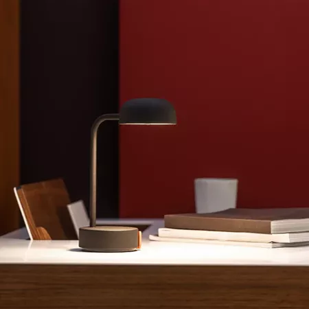 Darc — Une lampe design noire à bras articulé posée sur un bureau en bois clair, éclairant légèrement une pile de livres et un mug blanc devant un fon