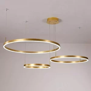 Darc — Trois suspensions lumineuses dorées de formes circulaires et ovales, suspendues à différentes hauteurs sur un mur blanc, créant une ambiance él