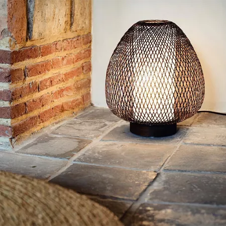 Darc — Une lampe design en forme de treillis métallique ajouré, posée sur une base noire, devant un mur en briques apparentes et un sol en carrelage g
