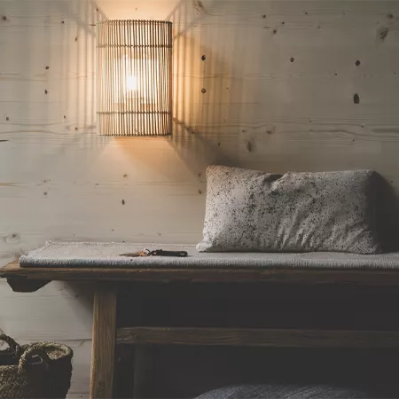 Darc — Une lampe murale en forme rectangulaire avec un abat-jour en rotin diffuse une lumière chaude sur un lit simple. Le lit est recouvert d’une cou