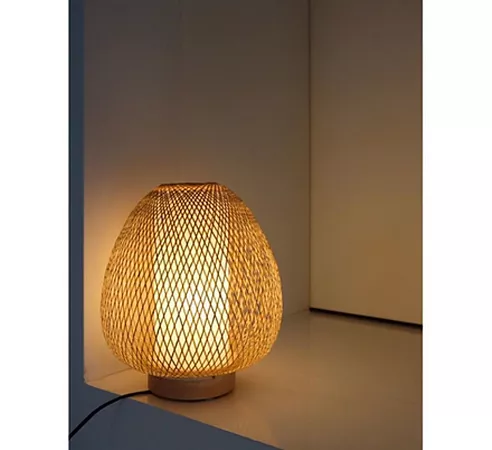 Darc — Lampe en forme ovale avec une structure en rotin tressé finement, posée sur un pied bas et éclairée par une lumière chaude dorée, créant une am