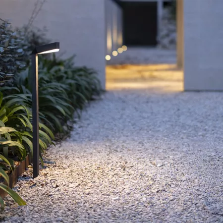Darc — Un projecteur LED moderne en métal noir éclairant une allée gravillonnée, entouré de plantes vertes et bordé d’une bordure en bois, avec une lu