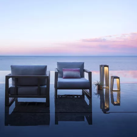 Darc — Deux fauteuils en bois et tissu gris, placés sur une terrasse en bois face à l'océan, avec un luminaire moderne à droite et un ciel dégradé de