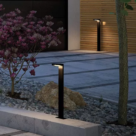 Darc — Deux lampadaires modernes en métal noir éclairant une allée de jardin pavée, entourés de plantes fleuries en fleurs roses et d’un petit arbre,
