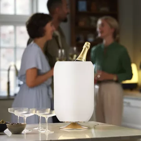 Darc — Un seau à champagne blanc design posé sur une table basse, avec une bouteille de champagne insérée et trois verres à pied à proximité. En arriè