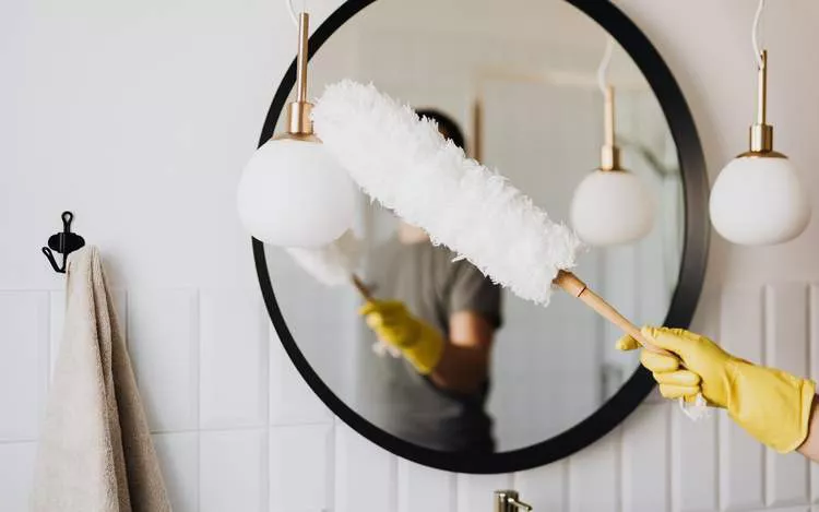 Darc — Une personne en gants jaunes nettoie un miroir rond encadré dans une salle de bain moderne, avec des luminaires blancs suspendus et une serviet