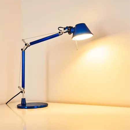 Darc — Lampe de bureau en métal bleu avec bras articulé et tête orientable, éclairant un mur blanc avec une lumière douce et chaude.