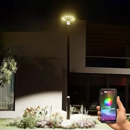 Darc — Une main tient un smartphone affichant un cercle coloré pour contrôler une lampe extérieure connectée, éclairant un jardin devant une maison mo