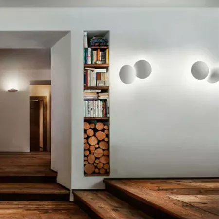 Darc — Une bibliothèque murale blanche intégrée dans un couloir clair, remplie de livres variés et de bûches empilées sur une étagère basse, éclairée