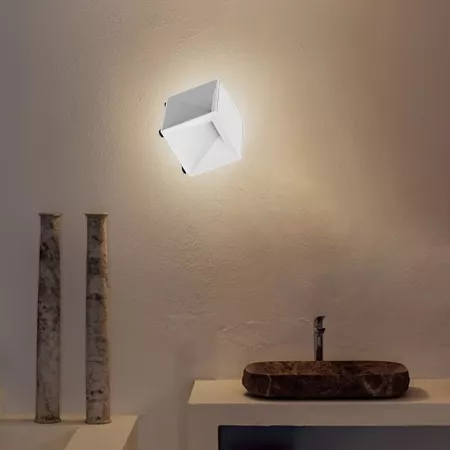 Darc — Une applique murale géométrique en métal blanc diffuse une lumière douce sur un mur clair. À gauche, deux colonnes verticales en pierre brute e