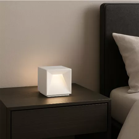 Darc — Une lampe cube blanche à lumière douce posée sur une table de chevet près d'un lit avec une tête de lit grise et un coussin beige, créant une a