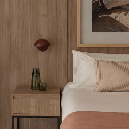 Darc — Chambre épurée avec un lit fait d’un drap blanc et d’un coussin à rayures horizontales beiges. À côté, une table de chevet en bois clair suppor