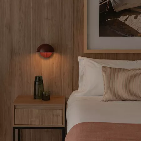 Darc — Une chambre sobre avec un lit bien fait, des coussins beige et blancs, une table de nuit en bois clair éclairée par une lampe design rouge et u