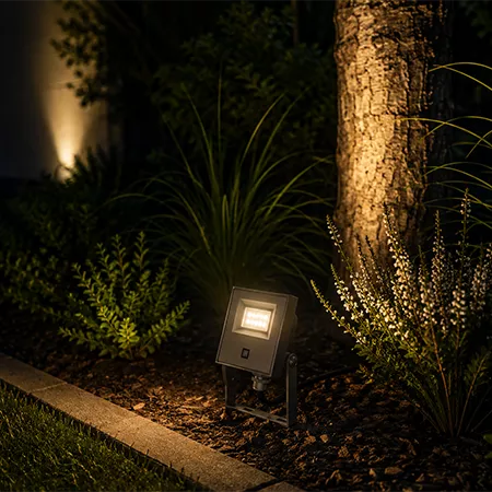 Darc — Un projecteur LED fixe posé sur un support métallique au sol, éclairant une zone de jardin avec des plantes et un arbre en arrière-plan, dans u