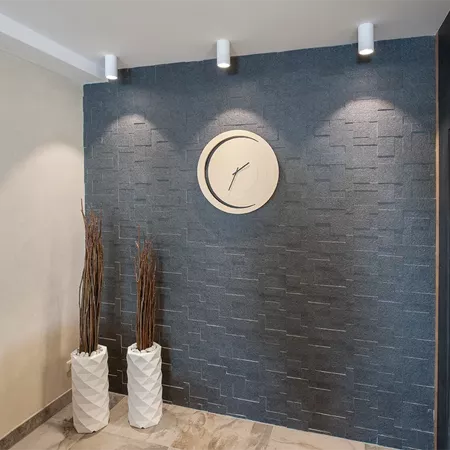 Darc — Un mur bleu imitation brique avec une horloge ronde blanche au centre, encadrée par trois spots lumineux. Deux pots en tissu blanc contenant de